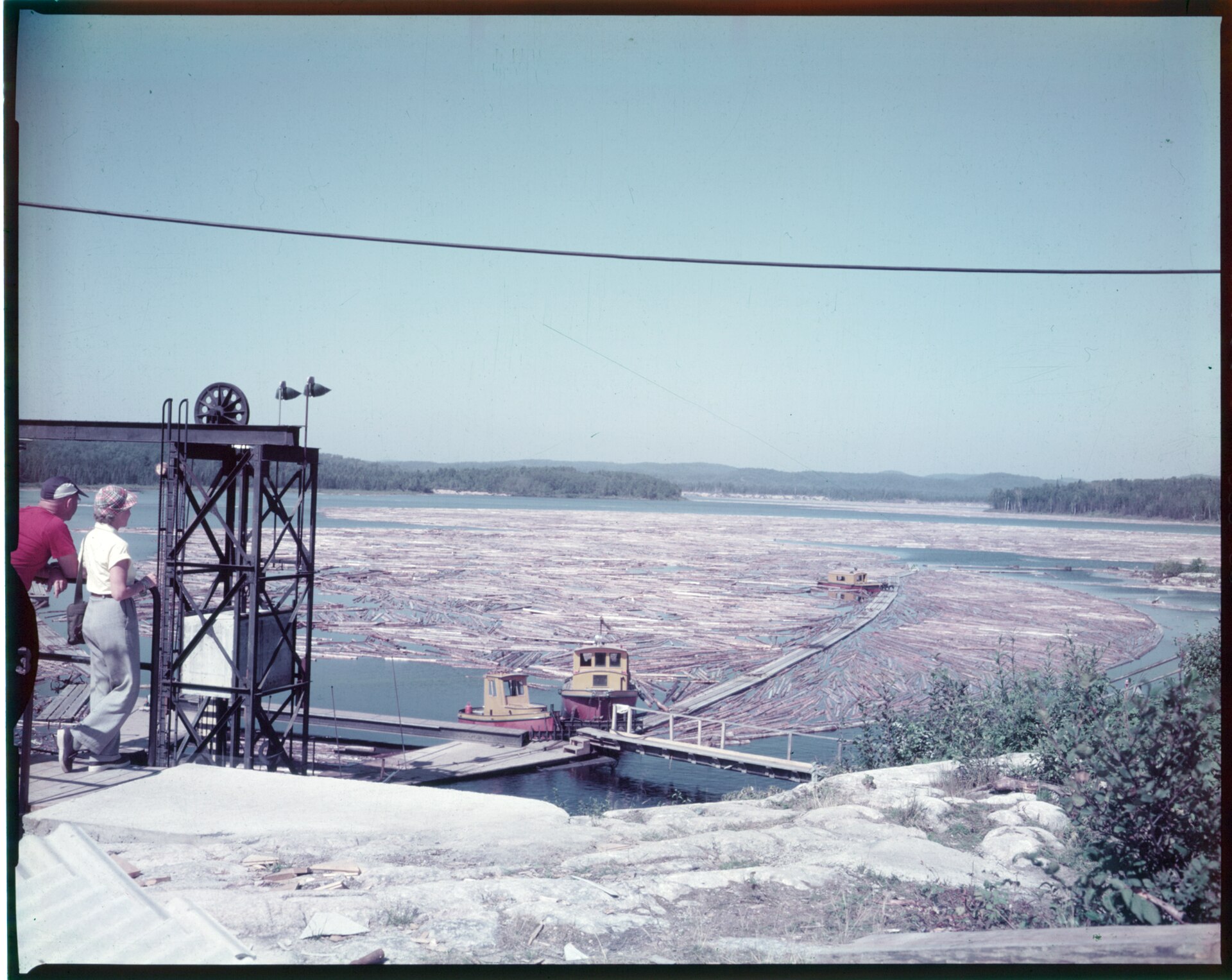 Archival colour photograph of a large boom of floating pulpwood logs
                covering a bay at Terrace Bay, Ontario, with two tugboats alongside
                and two people watching from a dock.