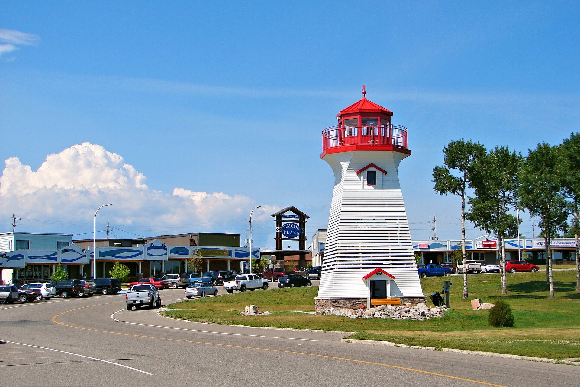 The main street of Terrace Bay, Ontario, with a white and red
                lighthouse-shaped lookout tower beside a row of low storefronts
                under a clear sky.
